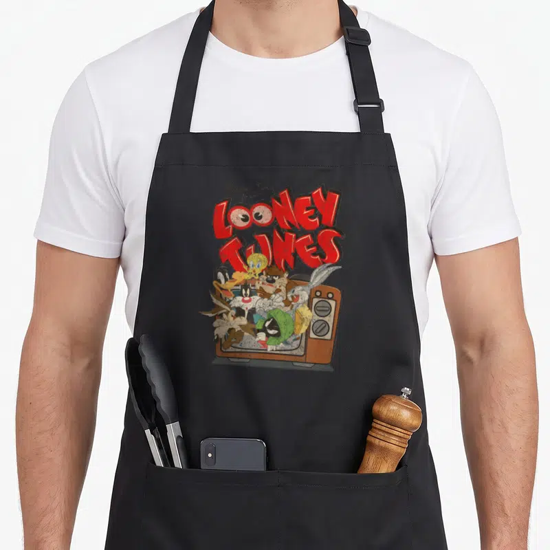 Funny cartoon saturday morning apron - Looney Tunes characters emerging from vintage TV with distressed red title text — shown on a black cotton canvas apron against a white background