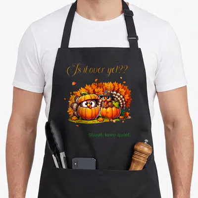 Thanksgiving gathering apron - Two cartoon turkeys hiding in orange pumpkins surrounded by brown and green leaves — shown on a black cotton canvas apron against a white background