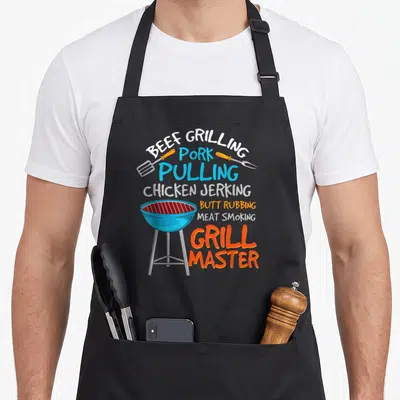 Beef grill master apron - Cartoon grill with stacked BBQ pun text in orange blue white red — shown on a black cotton canvas apron against a white background