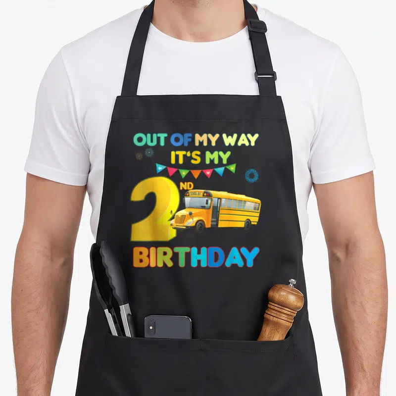 Bus 2nd apron - Cartoon school bus over yellow number 2 with Out of My Way It's My 2nd Birthday text and bunting flags — shown on a black cotton canvas apron against a white background