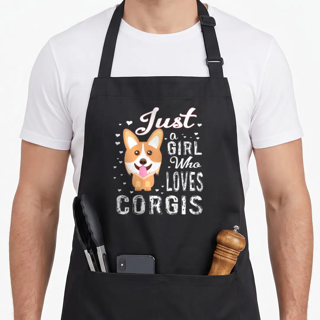 Corgis lvgteam apron - Cartoon corgi with pink tongue surrounded by text Just a GIRL Who LOVES CORVIS and scattered — shown on a black cotton canvas apron against a white background