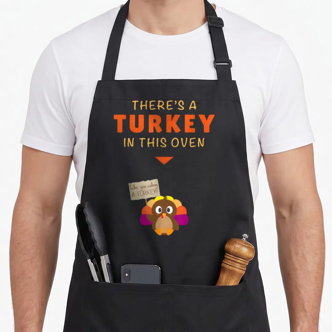 Thanksgiving funny turkey apron - Cartoon turkey holding a sign reading Who you calling A turkey under stacked text — shown on a black cotton canvas apron against a white background