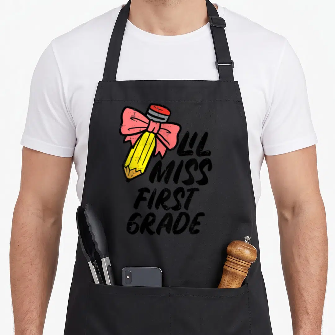 Lil miss grade apron - Yellow pencil with pink bow and L'L MISS FIRST GRADE text in distressed black — shown on a black cotton canvas apron against a white background