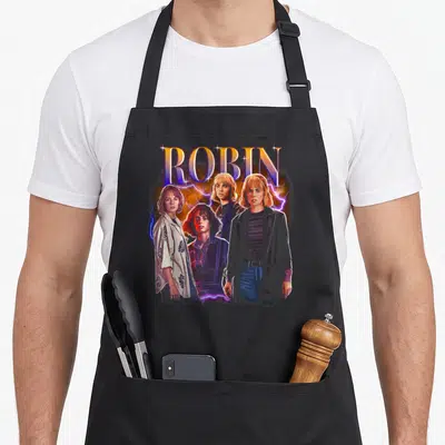 Heartthrob robin apron - Arched ROBIN text above four figures and lightning bolts in retro colors — shown on a black cotton canvas apron against a white background