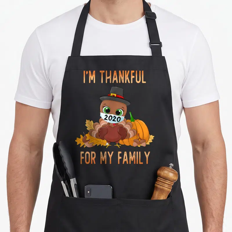 Family thankful apron - Cartoon turkey wearing 2020 mask and pilgrim hat next to pumpkin and leaves — shown on a black cotton canvas apron against a white background
