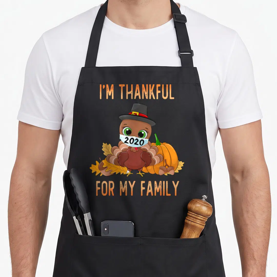 Family thankful apron - Cartoon turkey wearing 2020 mask and pilgrim hat next to pumpkin and leaves — shown on a black cotton canvas apron against a white background