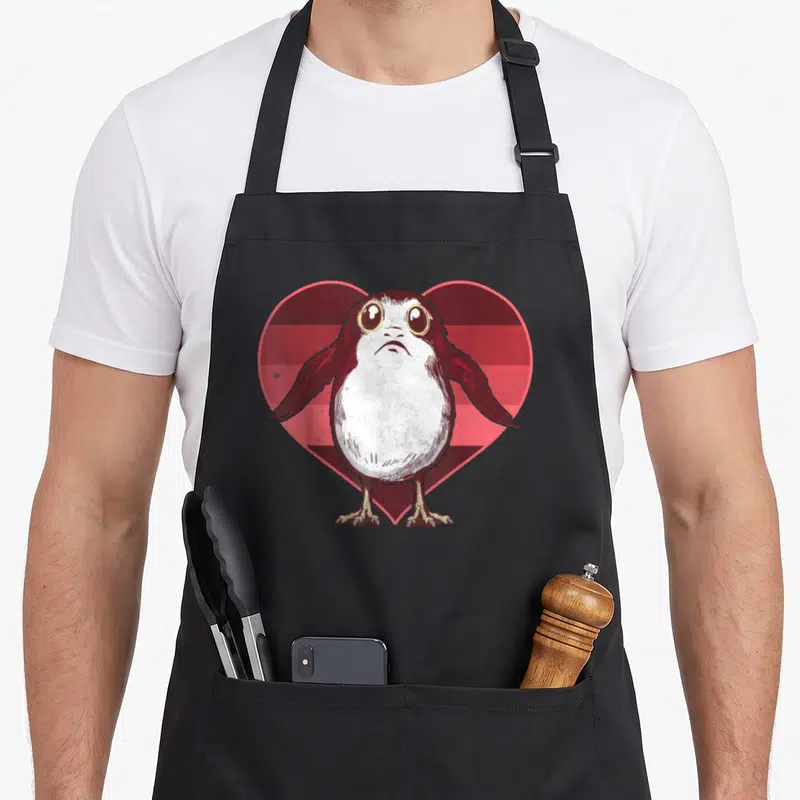 Porg valentine apron - Cartoon penguin standing in front of a heart with red and pink horizontal stripes — shown on a black cotton canvas apron against a white background