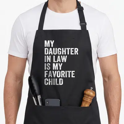 Funny father mom and daughter aprons - White stacked text reading My Daughter In Law Is My Favorite Child on black — shown on a black cotton canvas apron against a white background