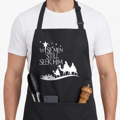Seek christmas apron - White text Wise Men Still Seek Him above three camel riders heading to a city skyline — shown on a black cotton canvas apron against a white background