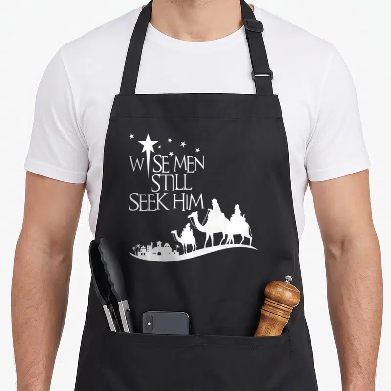 Seek christmas apron - White text Wise Men Still Seek Him above three camel riders heading to a city skyline — shown on a black cotton canvas apron against a white background