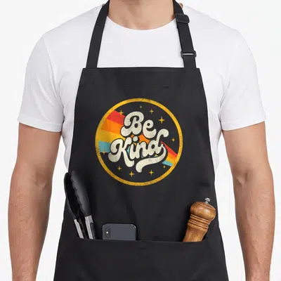 Kind anti bullying awareness co apron - Retro Be Kind text in yellow circle with red orange and blue diagonal stripes — shown on a black cotton canvas apron against a white background
