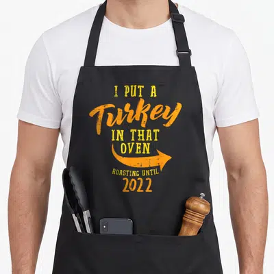 Oven pregnancy thanksgiving apron - Stacked text I Put A Turkey In That Oven Roasting Until 2022 with orange arrow — shown on a black cotton canvas apron against a white background