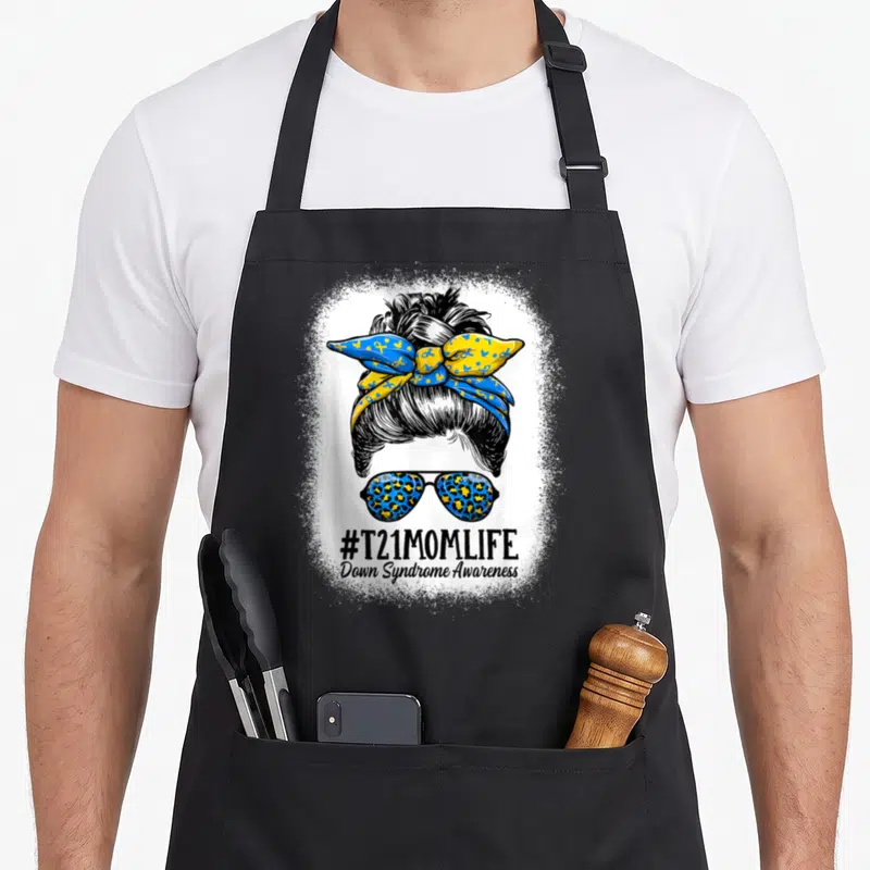 Syndrome messy aprons - Cartoon woman bun with blue yellow ribbon sunglasses and #T21MOMLIFE text — shown on a black cotton canvas apron against a white background