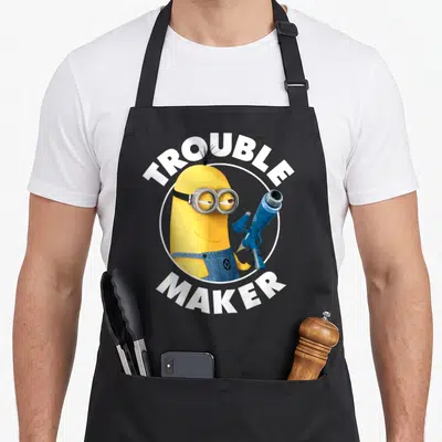 Despicable maker apron - Yellow minion in white circle with trouble maker text arching above and below — shown on a black cotton canvas apron against a white background