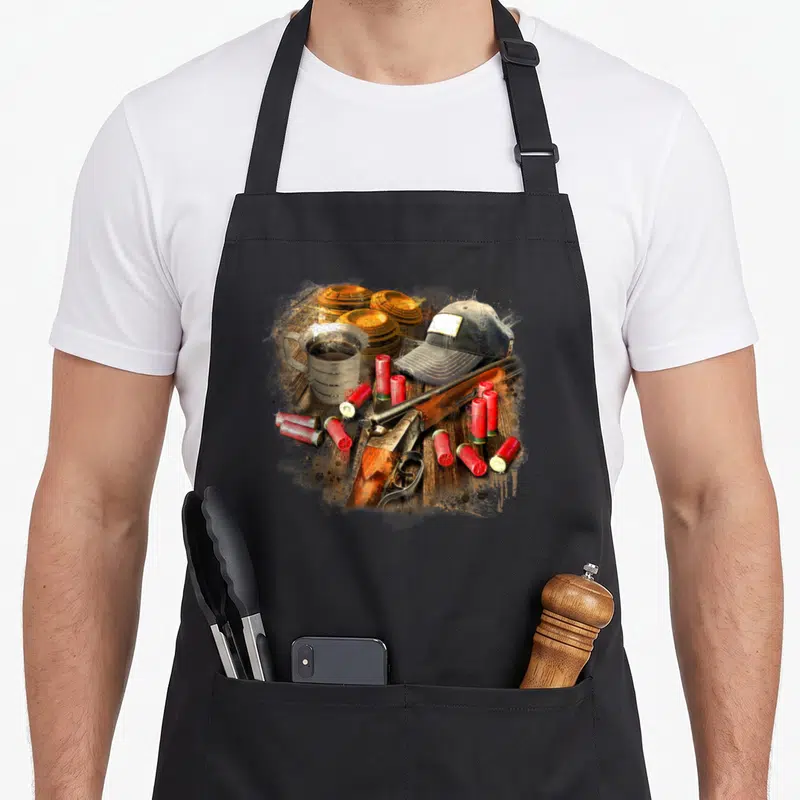 Hat clay apron - Realistic shotgun, red shells, metal cup, and clay pigeons on a wooden table — shown on a black cotton canvas apron against a white background