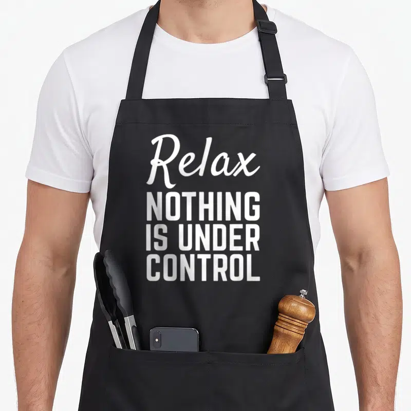 Relax meditation apron - White stacked text reading Relax NOTHING IS UNDER CONTROL on a dark background — shown on a black cotton canvas apron against a white background