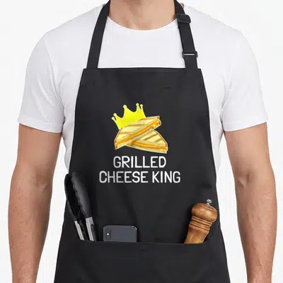 Cheese king apron - Cartoon grilled cheese sandwich with yellow crown and white text reading GRILLED CHEESE KING — shown on a black cotton canvas apron against a white background