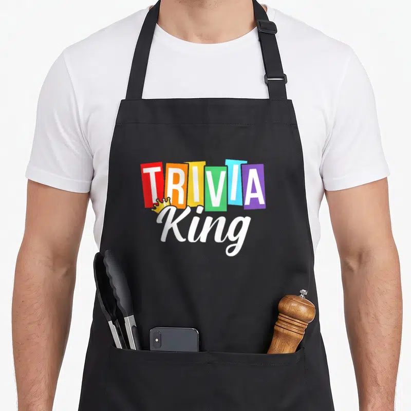 Trivia king apron - TRIVIA in colored blocks with King script and yellow crown graphic — shown on a black cotton canvas apron against a white background
