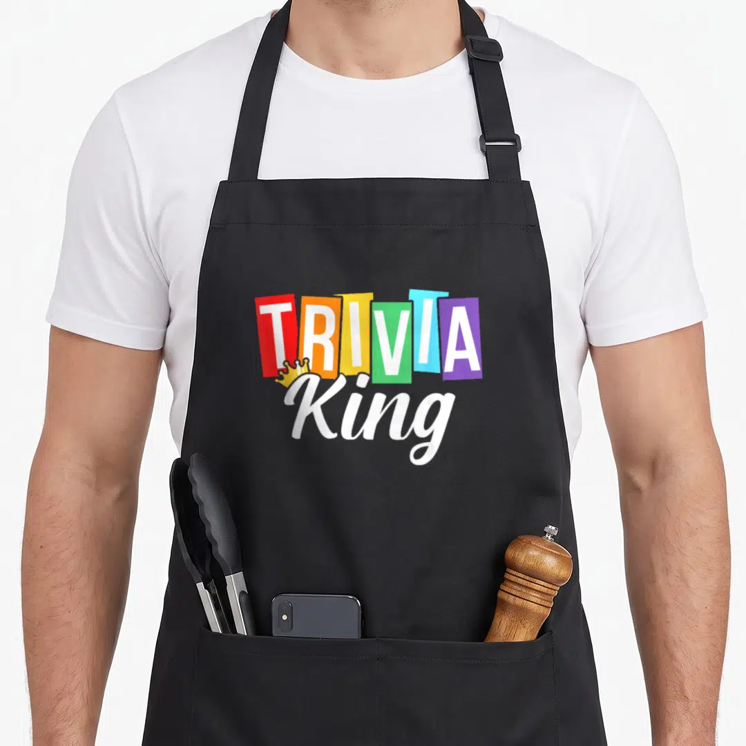 Trivia king apron - TRIVIA in colored blocks with King script and yellow crown graphic — shown on a black cotton canvas apron against a white background