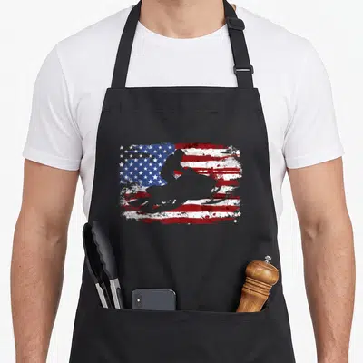 Funny snowmobile american flag apron - Grey snowmobile silhouette over distressed red white and blue flag — shown on a black cotton canvas apron against a white background