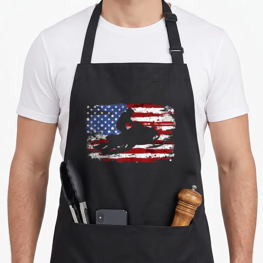 Funny snowmobile american flag apron - Grey snowmobile silhouette over distressed red white and blue flag — shown on a black cotton canvas apron against a white background