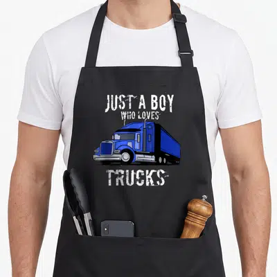 Funny semi truck apron - Blue cartoon semi-truck with white text JUST A BOY WHO LOVES TRUCKS on grey background — shown on a black cotton canvas apron against a white background