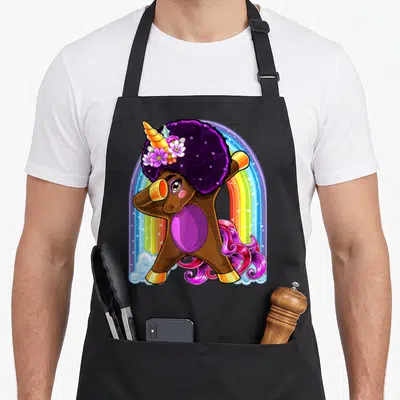 Dabbing african american aprons - Brown cartoon unicorn with purple afro, pink mane, flowers, and rainbow background — shown on a black cotton canvas apron against a white background