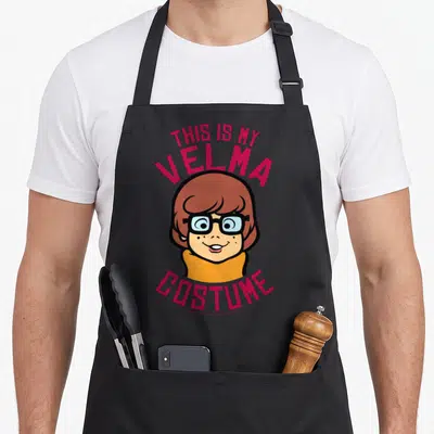 Velma costume comfort apron - Cartoon Velma head with pink text THIS IS MY VELMA COSTUME arched above and below — shown on a black cotton canvas apron against a white background