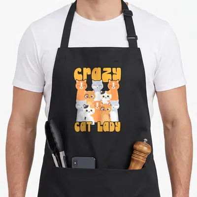 Kitten animal crazy cat lady apron - Nine cartoon cats in grey orange and white sandwiched between yellow crazy and CAT — shown on a black cotton canvas apron against a white background