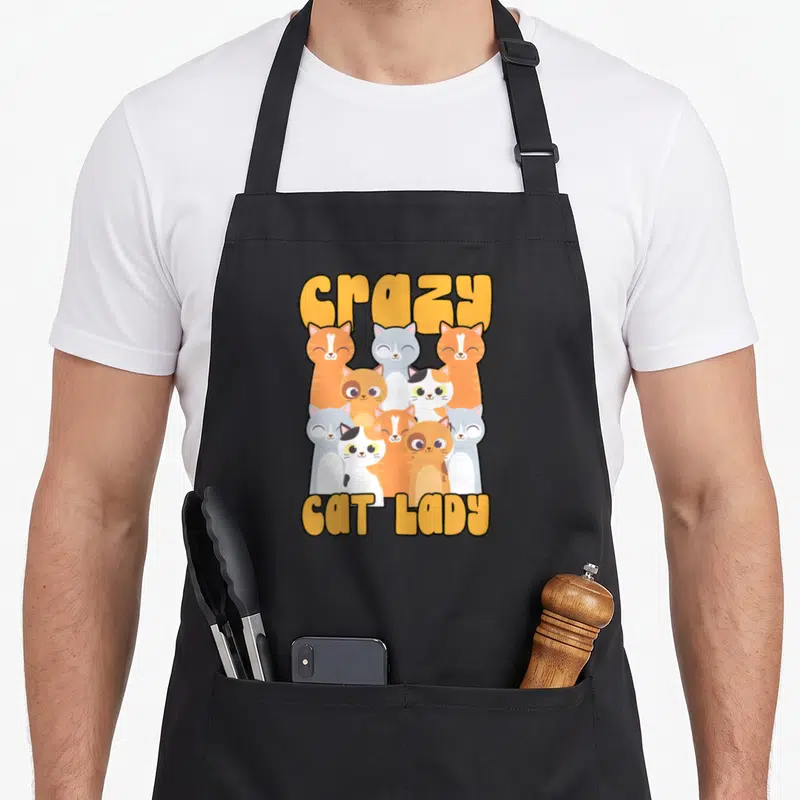 Kitten animal crazy cat lady apron - Nine cartoon cats in grey orange and white sandwiched between yellow crazy and CAT — shown on a black cotton canvas apron against a white background