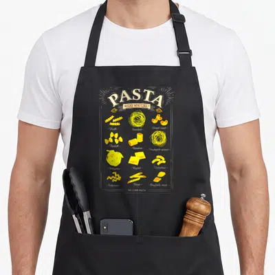 Funny italian pasta apron - Yellow typography chart of twelve pasta shapes with names like Fusilli, Spaghetti — shown on a black cotton canvas apron against a white background