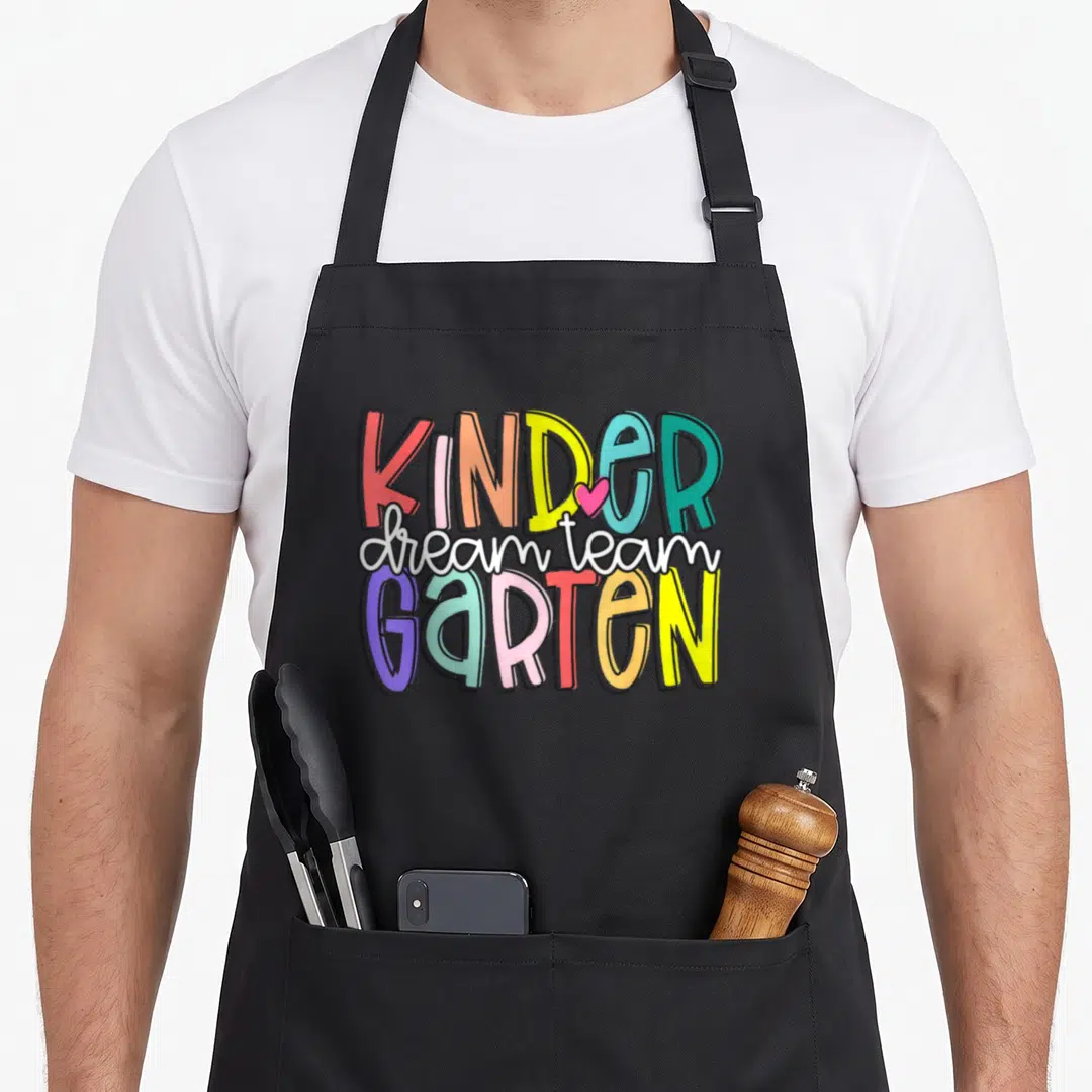 Dream kindergarten teacher apron - Stacked multicolor text reading KINDER dream team GARTEN with a pink heart between K — shown on a black cotton canvas apron against a white background