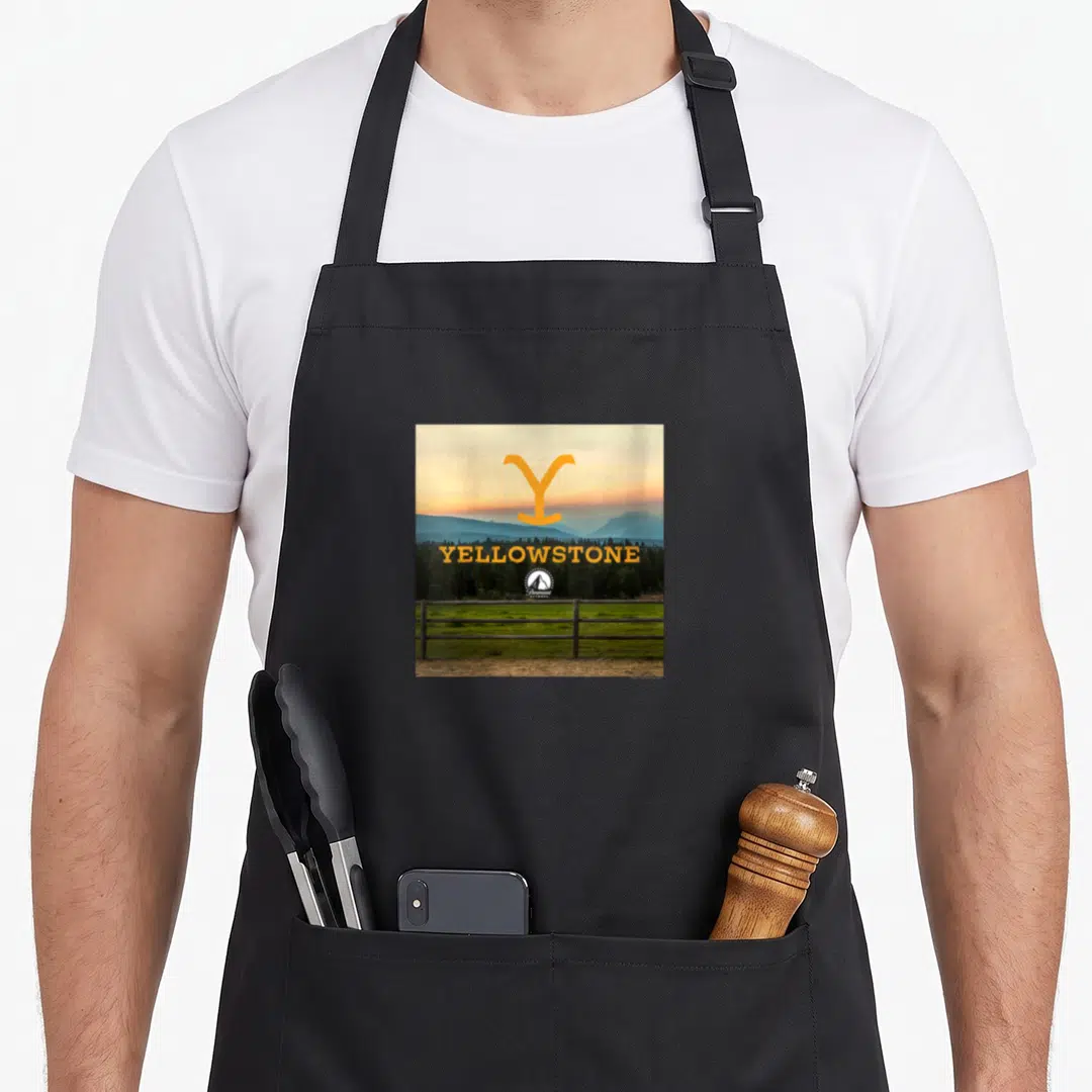 Dutton country apron - Yellow Y symbol above YELLOWSTONE text over mountain photo with wooden fence and small tent logo — shown on a black cotton canvas apron against a white background