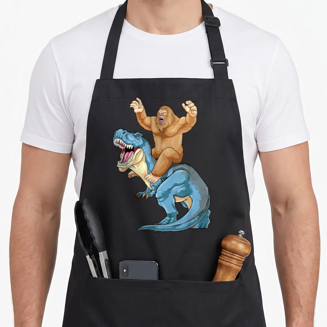 Sasquatch dinosaur apron - Cartoon brown Bigfoot riding a blue T-Rex facing left with open mouth — shown on a black cotton canvas apron against a white background