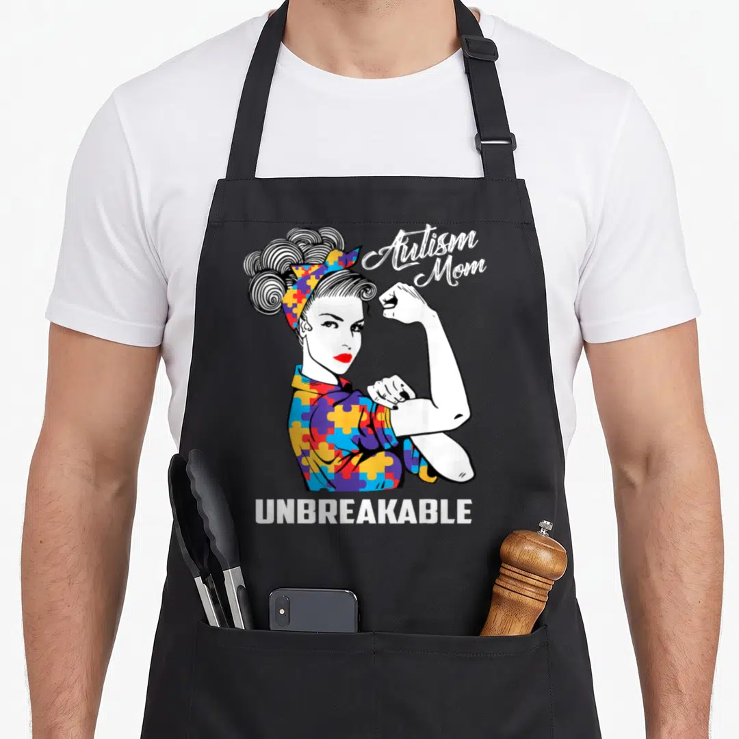 Autism mama apron - Cartoon woman flexing arm wearing blue red yellow purple puzzle pieces with Autism Mom text — shown on a black cotton canvas apron against a white background