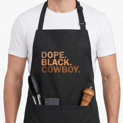Dope cowboy apron - Stacked distressed orange and brown text reading DOPE. BLACK. COWBOY — shown on a black cotton canvas apron against a white background