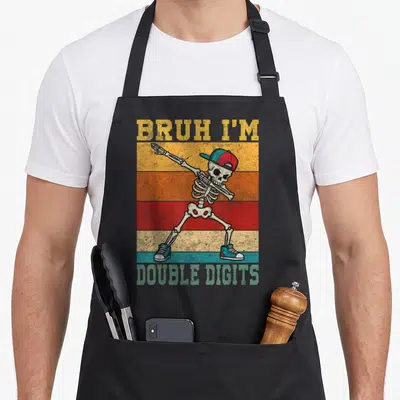 Cool bruh birthday apron - Dabbing skeleton wearing cap and sneakers over retro striped background with distressed text — shown on a black cotton canvas apron against a white background