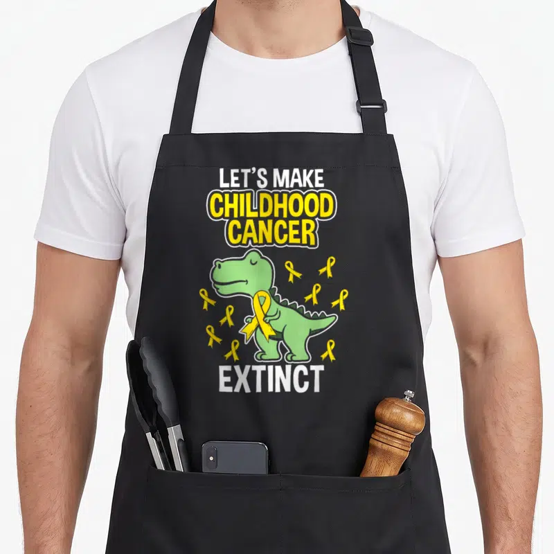 Childhood dinosaur apron - Green cartoon dinosaur holding yellow ribbon with Let's Make Childhood Cancer Extinct text — shown on a black cotton canvas apron against a white background