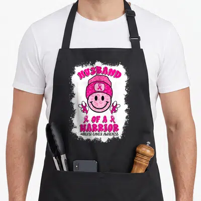 Funny husband breast cancer awareness aprons - Pink leopard smiley face wearing beanie with HUSBAND OF A WARRIOR text — shown on a black cotton canvas apron against a white background