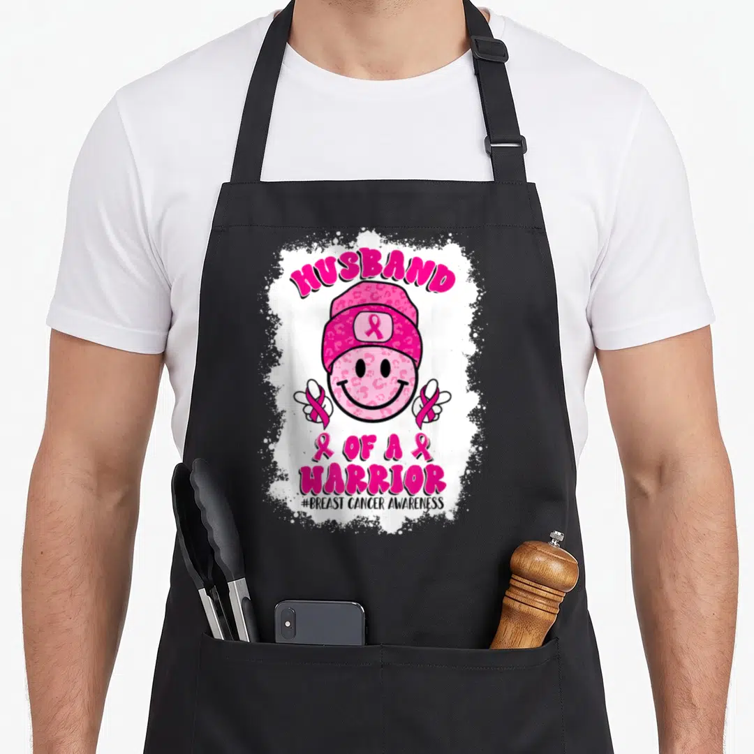 Funny husband breast cancer awareness aprons - Pink leopard smiley face wearing beanie with HUSBAND OF A WARRIOR text — shown on a black cotton canvas apron against a white background