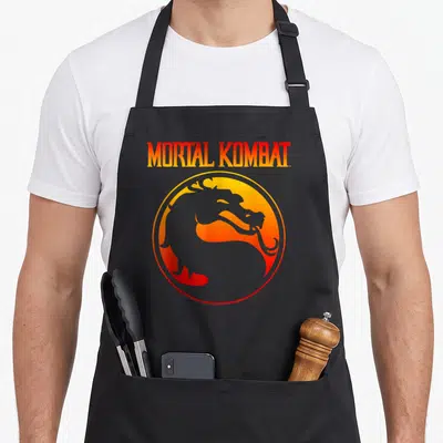 Klassic video game apron - MORTAL KOMBAT gradient text above circular orange yellow grey dragon silhouette emblem — shown on a black cotton canvas apron against a white background