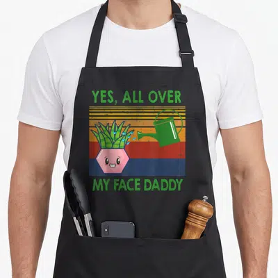 Face daddy apron - Yes All Over My Face Daddy text with cartoon plant and watering can on striped background — shown on a black cotton canvas apron against a white background