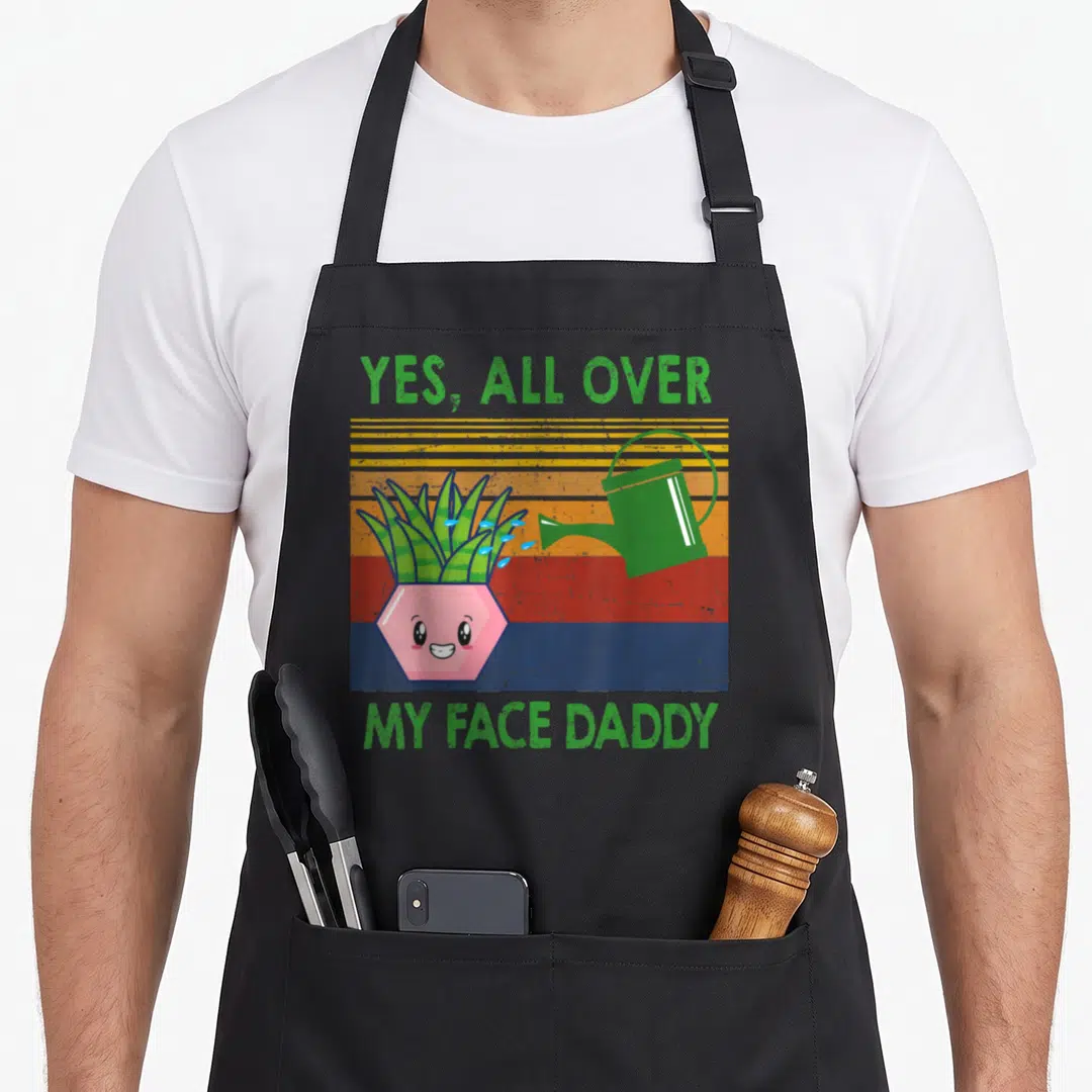 Face daddy apron - Yes All Over My Face Daddy text with cartoon plant and watering can on striped background — shown on a black cotton canvas apron against a white background