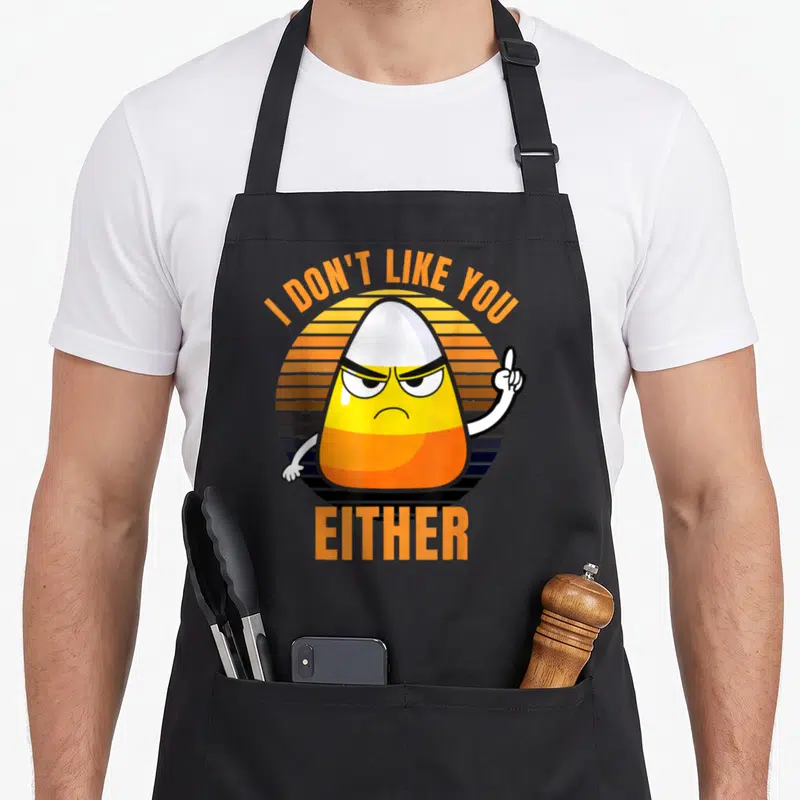 Either halloween apron - Angry candy corn with raised finger under arched text I DON'T LIKE YOU, EITHER on striped — shown on a black cotton canvas apron against a white background