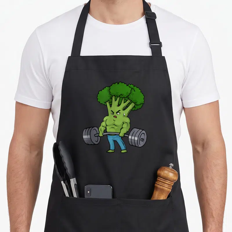 Deadlift bodybuilding apron - Cartoon muscular broccoli lifting grey barbell wearing blue pants — shown on a black cotton canvas apron against a white background