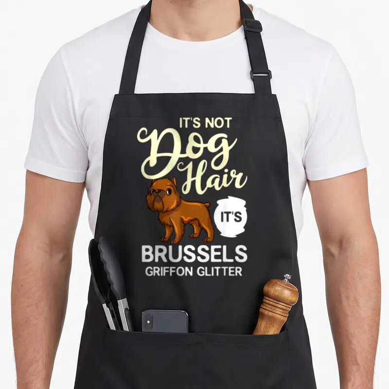 Griffon puppy dog apron - Cartoon brown Brussels Griffon dog with It's Not Dog Hair Brussels Griffon Glitter text — shown on a black cotton canvas apron against a white background