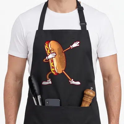 Dabbing hot dog apron - Cartoon hot dog with white gloves and shoes dancing in tan, red, and yellow — shown on a black cotton canvas apron against a white background
