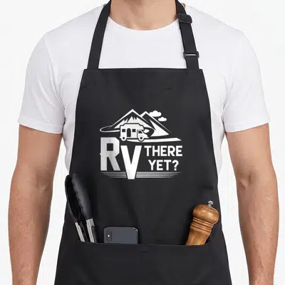 Roadtrip camping apron - White RV illustration on road with mountains and text RV THERE YET — shown on a black cotton canvas apron against a white background