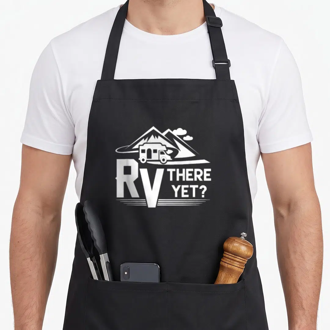 Roadtrip camping apron - White RV illustration on road with mountains and text RV THERE YET — shown on a black cotton canvas apron against a white background