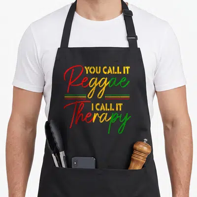 Call my therapy apron - Stacked red yellow and green text reading You Call It Reggae I Call It Therapy — shown on a black cotton canvas apron against a white background